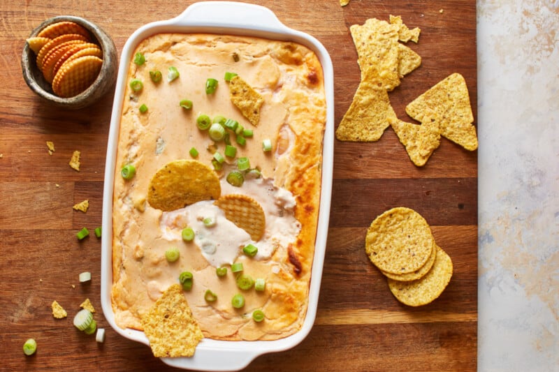 overhead view of shrimp dip in a white baking pan on a wooden cutting board surrounded by tortilla chips and crackers.