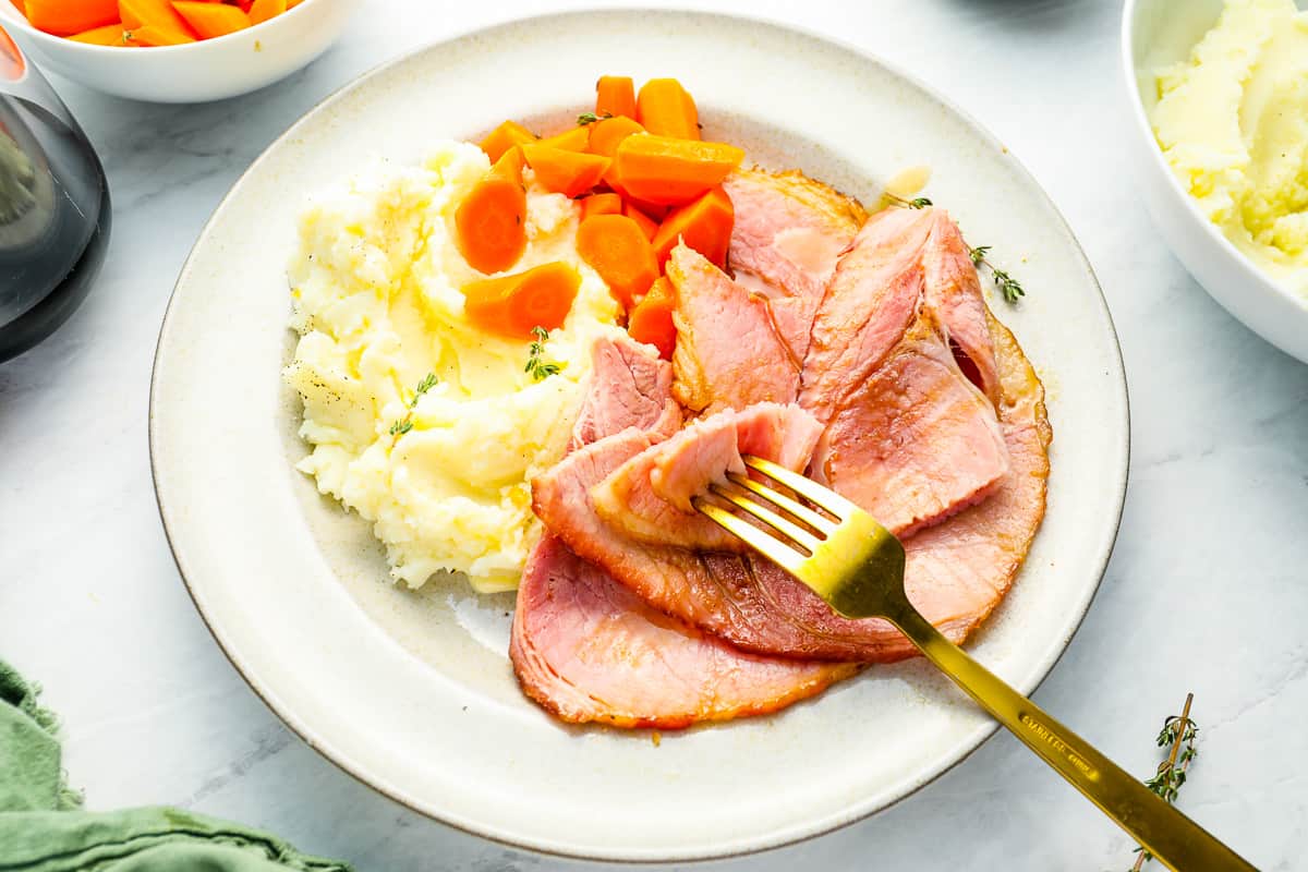 three-quarters view of a serving of smoked ham on a white plate with mashed potatoes, carrots, and a fork.