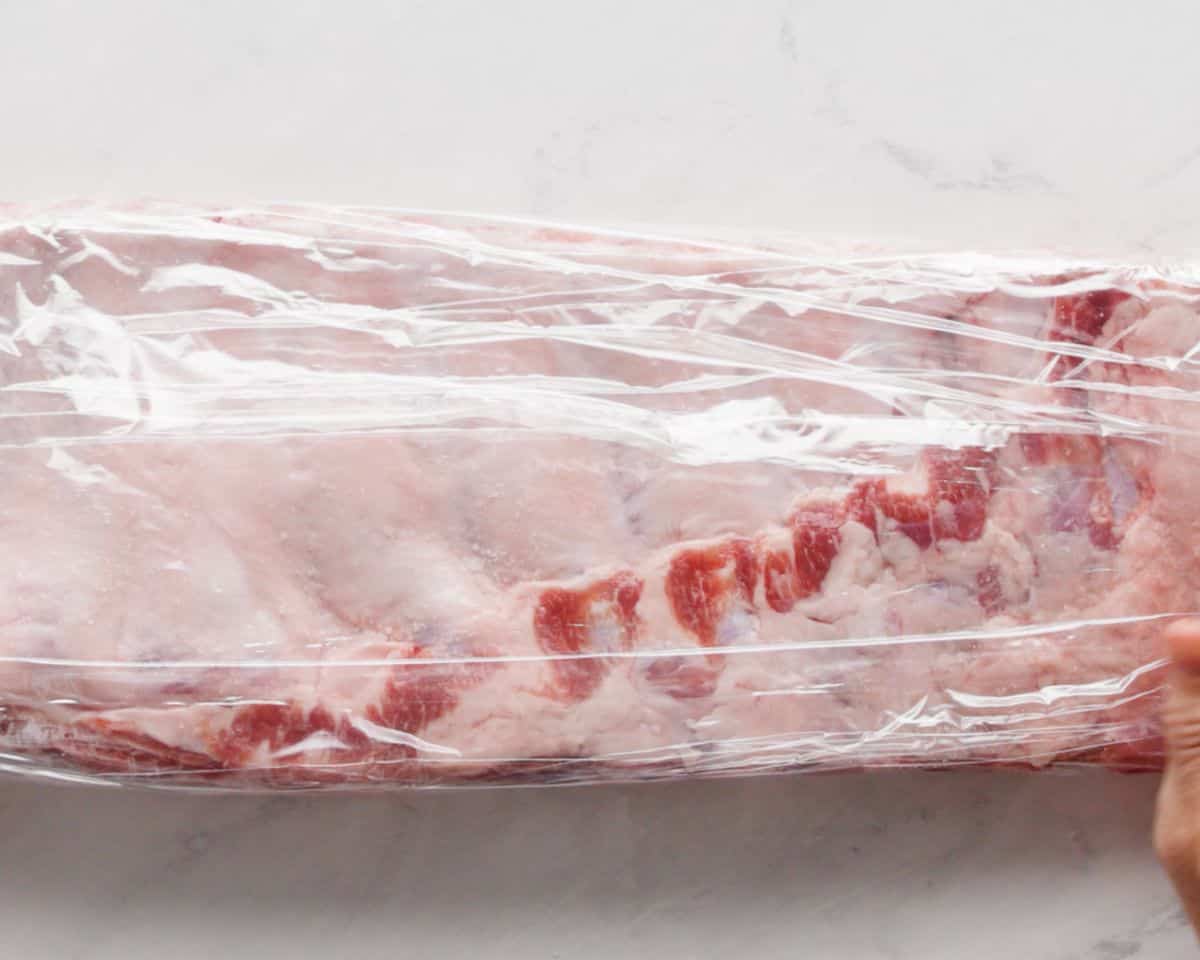 Raw St. Louis ribs wrapped tightly in plastic wrap during the dry brining process.