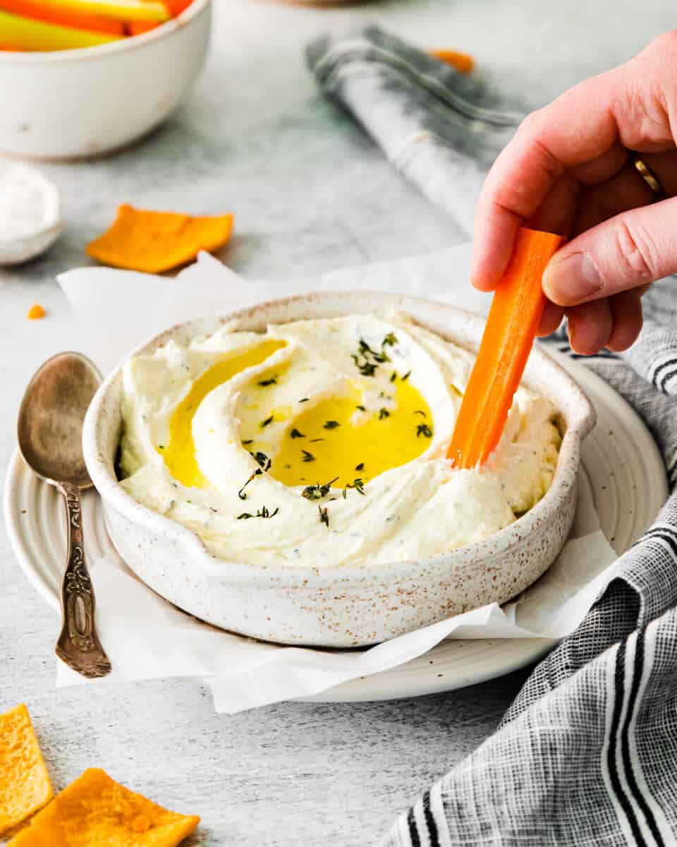 three-quarters view of a hand dipping a carrot stick into whipped feta dip with olive oil on top.