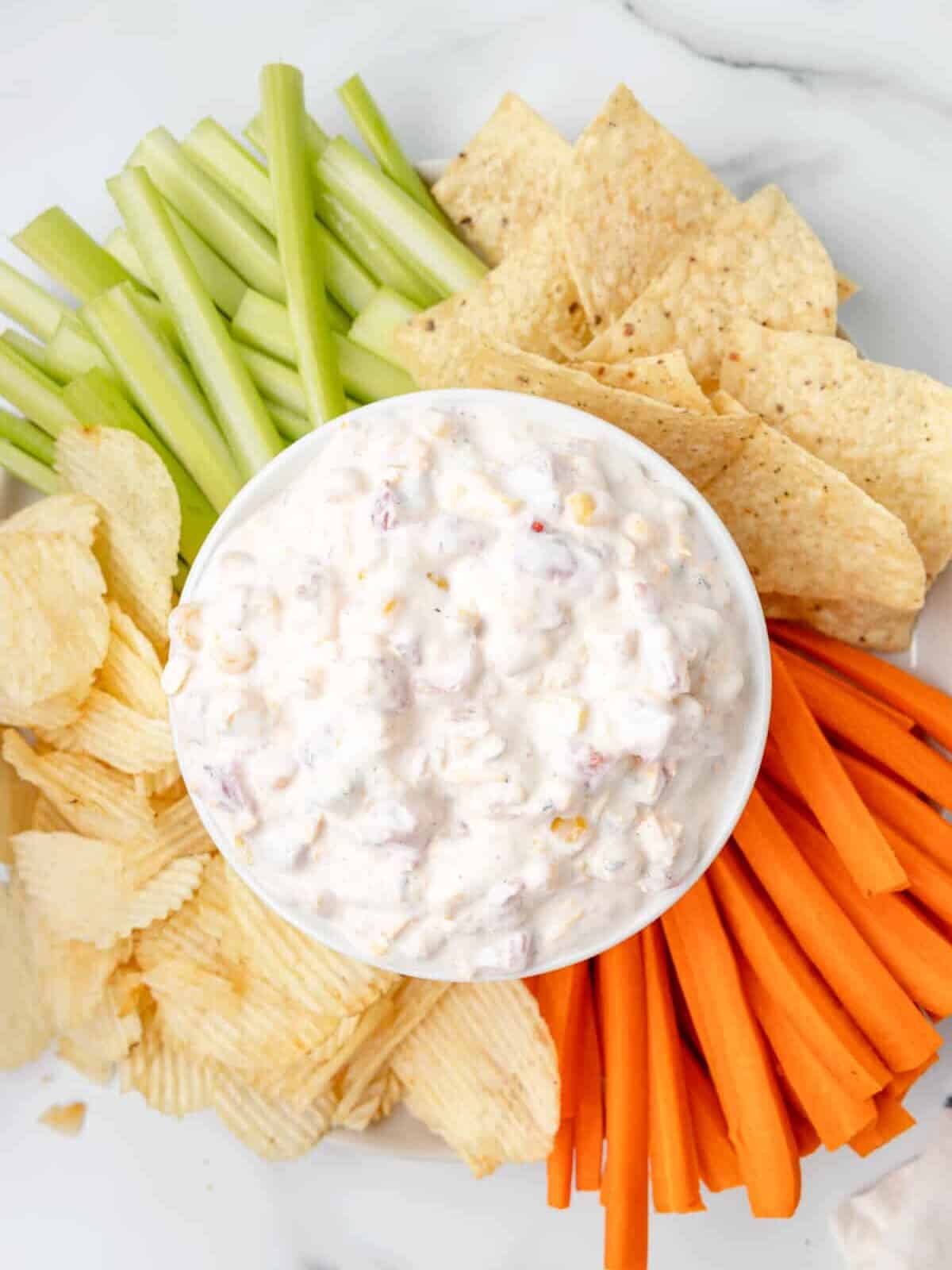 overhead view of fiesta ranch corn dip in a white bowl surrounded by wavy potato chips, carrot sticks, tortilla chips, and celery sticks.