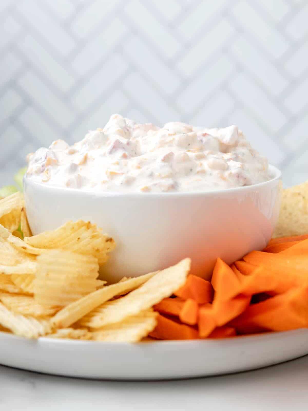 side view of cold fiesta ranch dip in a white bowl surrounded by wavy potato chips and carrot sticks,