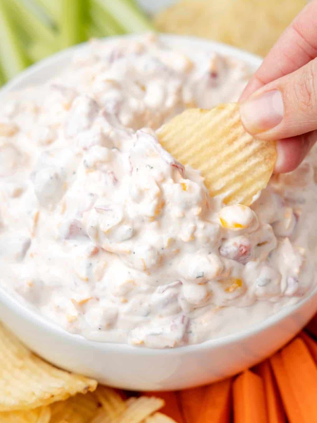 three-quarters view of a hand dipping a wavy potato chip into fiesta ranch dip in a white bowl surrounded by wavy potato chips, carrot sticks, tortilla chips, and celery sticks.