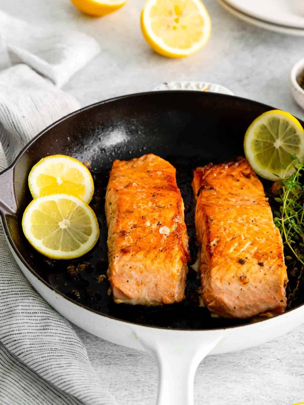 three-quarters view of 2 cooked salmon filets in a cast iron skillet with thyme and lemon.
