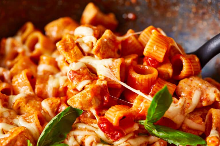 A close-up of a dish featuring rigatoni pasta mixed with chunks of chicken, covered in a rich tomato sauce, and topped with melted cheese. Fresh basil leaves garnish the dish, adding a touch of green.