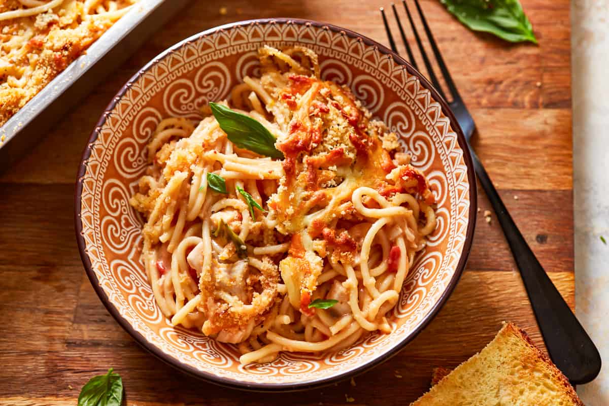 A bowl of chicken spaghetti garnished with basil on a wooden table with bread. 
