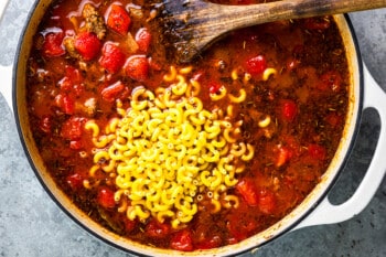 macaroni added to a pot full of goulash with a wooden spoon.