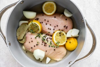 overhead view of 2 raw chicken breasts covered in aromatics and poaching liquid in a pot.