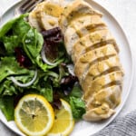 overhead view of a sliced poached chicken breast on a white plate with greens, lemon slices, and a fork.