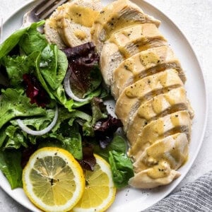 overhead view of a sliced poached chicken breast on a white plate with greens, lemon slices, and a fork.