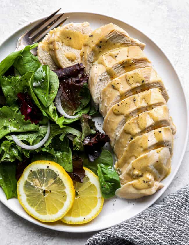 overhead view of a sliced poached chicken breast on a white plate with greens, lemon slices, and a fork.