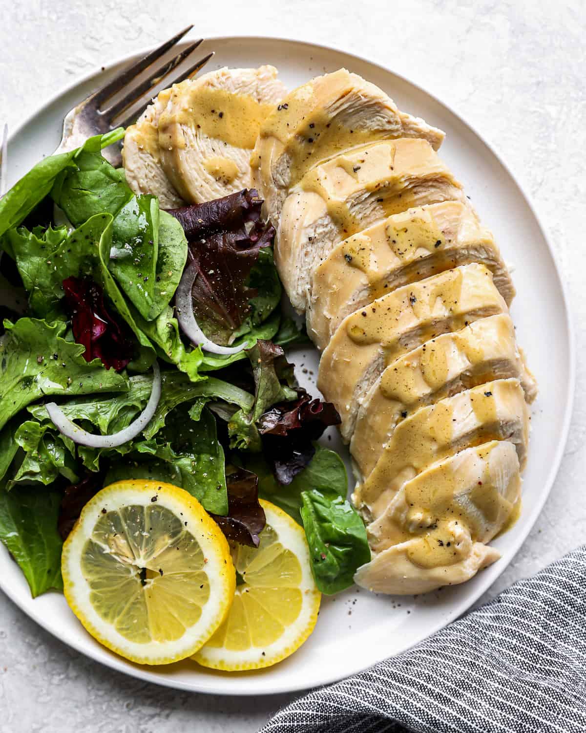 overhead view of a sliced poached chicken breast on a white plate with greens, lemon slices, and a fork.