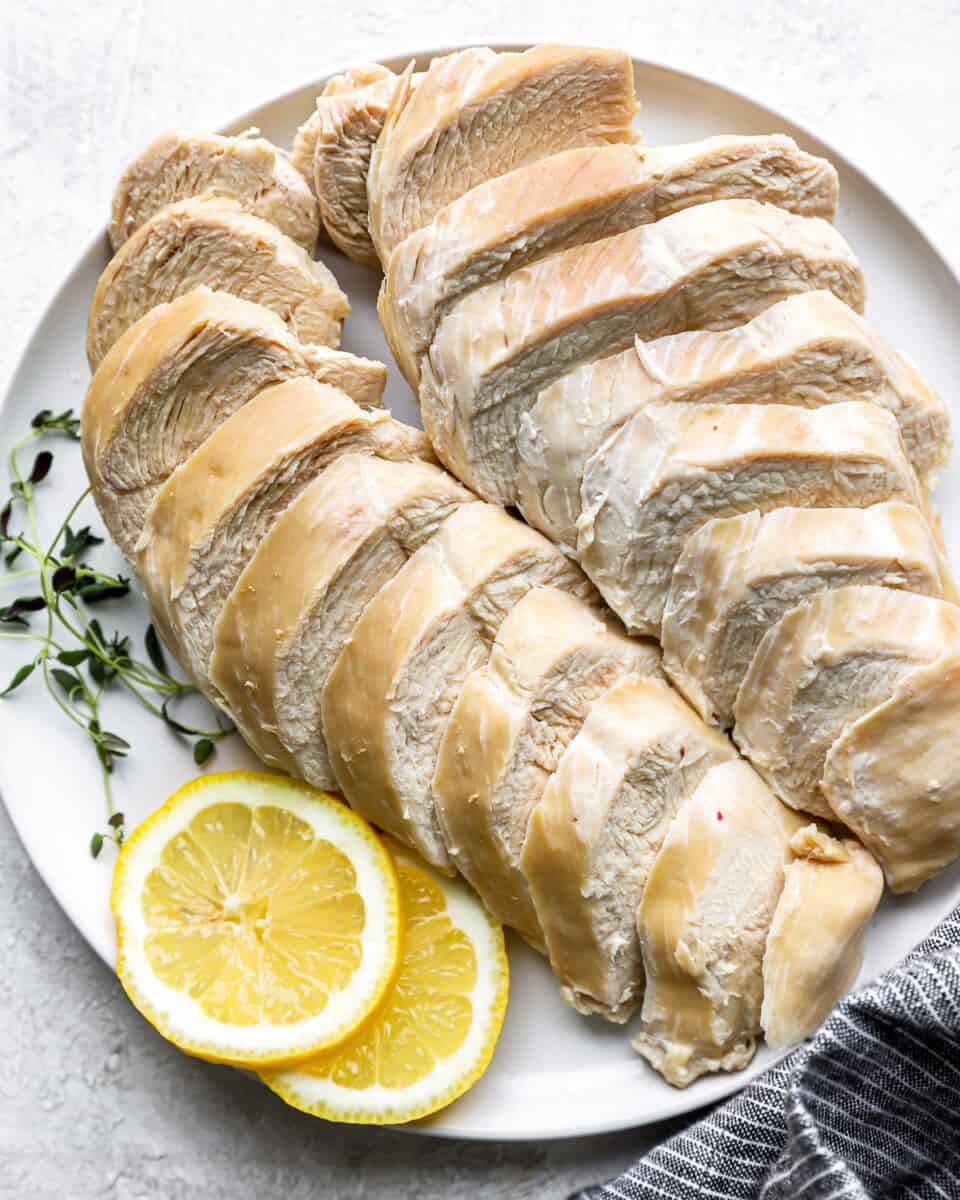 overhead view of 2 sliced poached chicken breasts on a white plate with lemon slices and thyme sprigs.