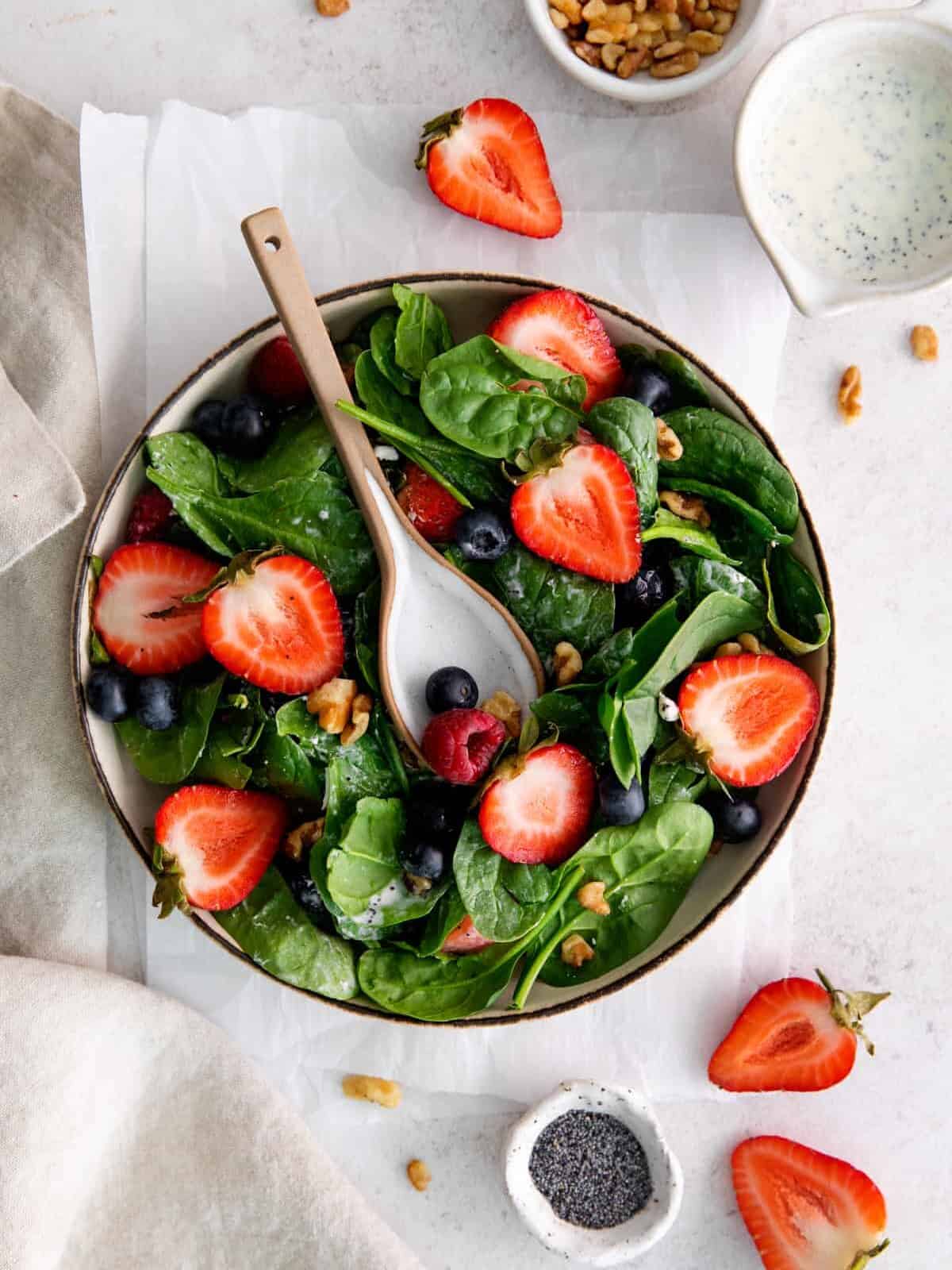 overhead view of spinach berry salad filled with strawberries, blueberries, raspberries, walnuts, and goat cheese, in a beige bowl with a serving spoon.