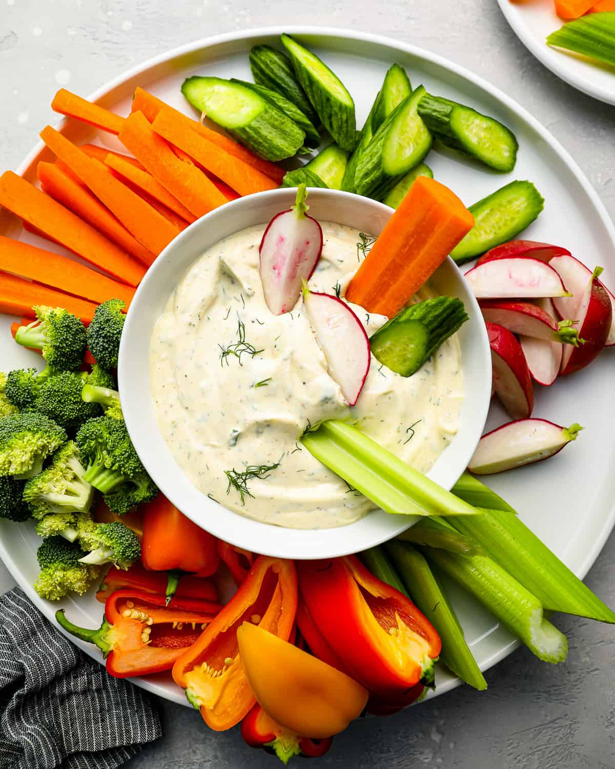 a vegetable tray filled with carrots, broccoli, peppers, celery, and radishes; with a bowl of veggie dip at the center, with vegetables partially dipped into it.
