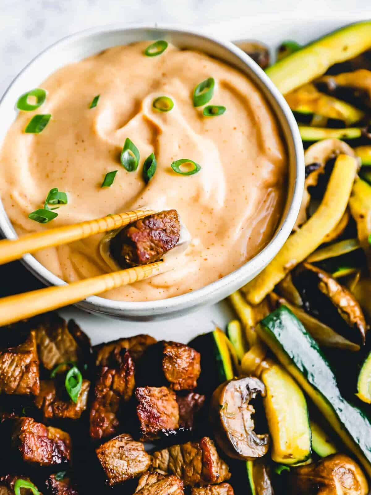 three-quarters view of a pair of chopsticks dipping a cube of steak into DIY yum yum sauce in a white bowl.