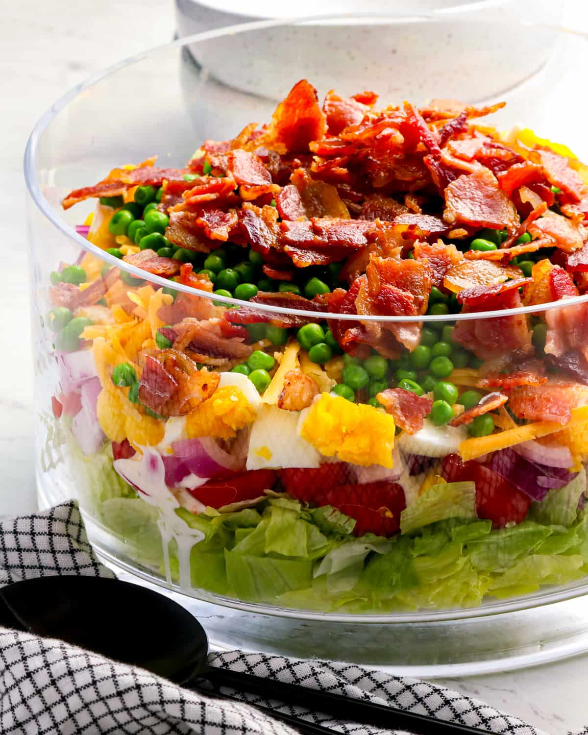 A 7 layer salad with bacon, peas, lettuce, onions, tomatoes, cheese, and eggs in a bowl.