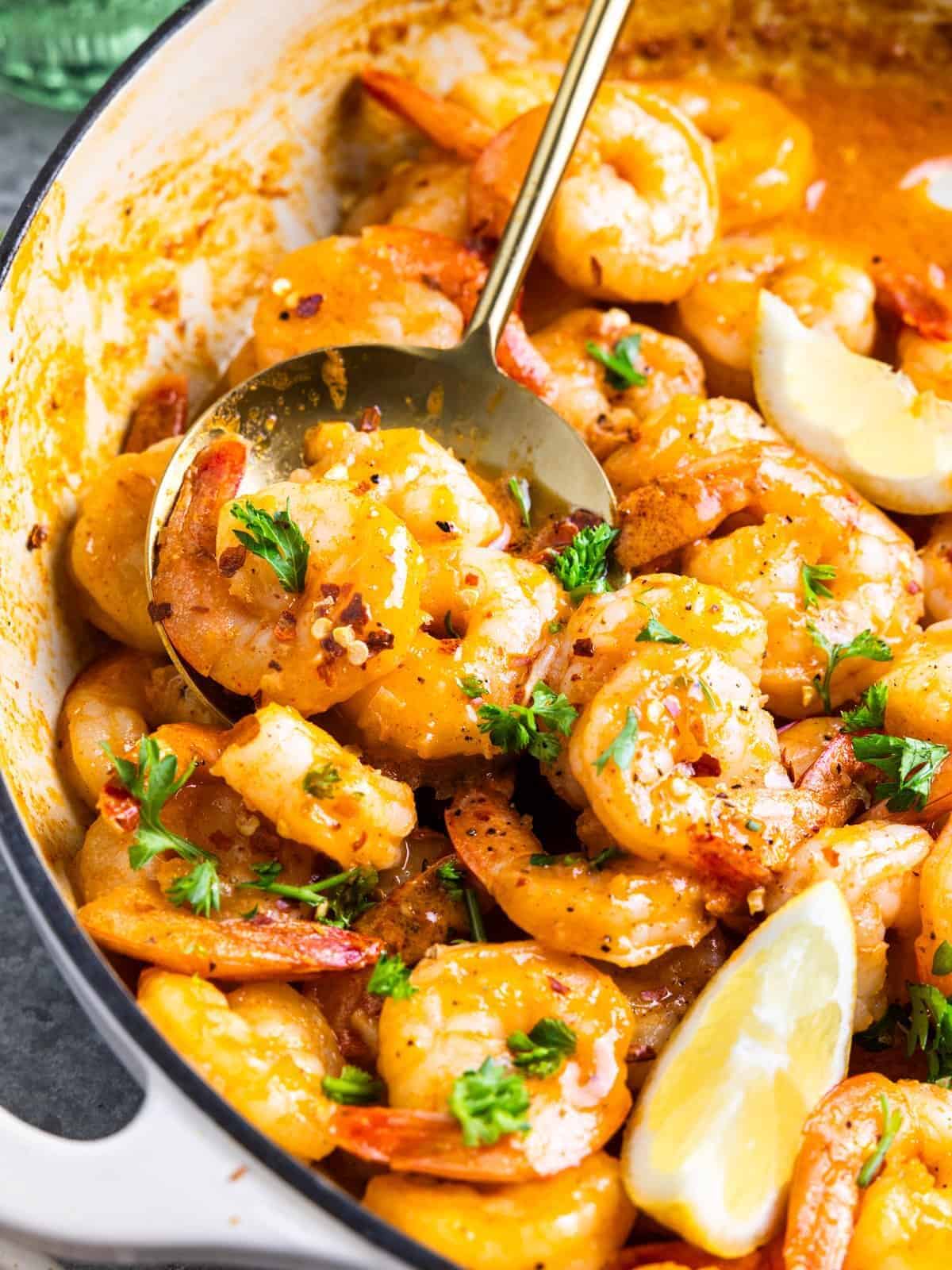 close-up partial three-quarters view of garlic butter shrimp in a white pan with a spoon and lemon wedges.