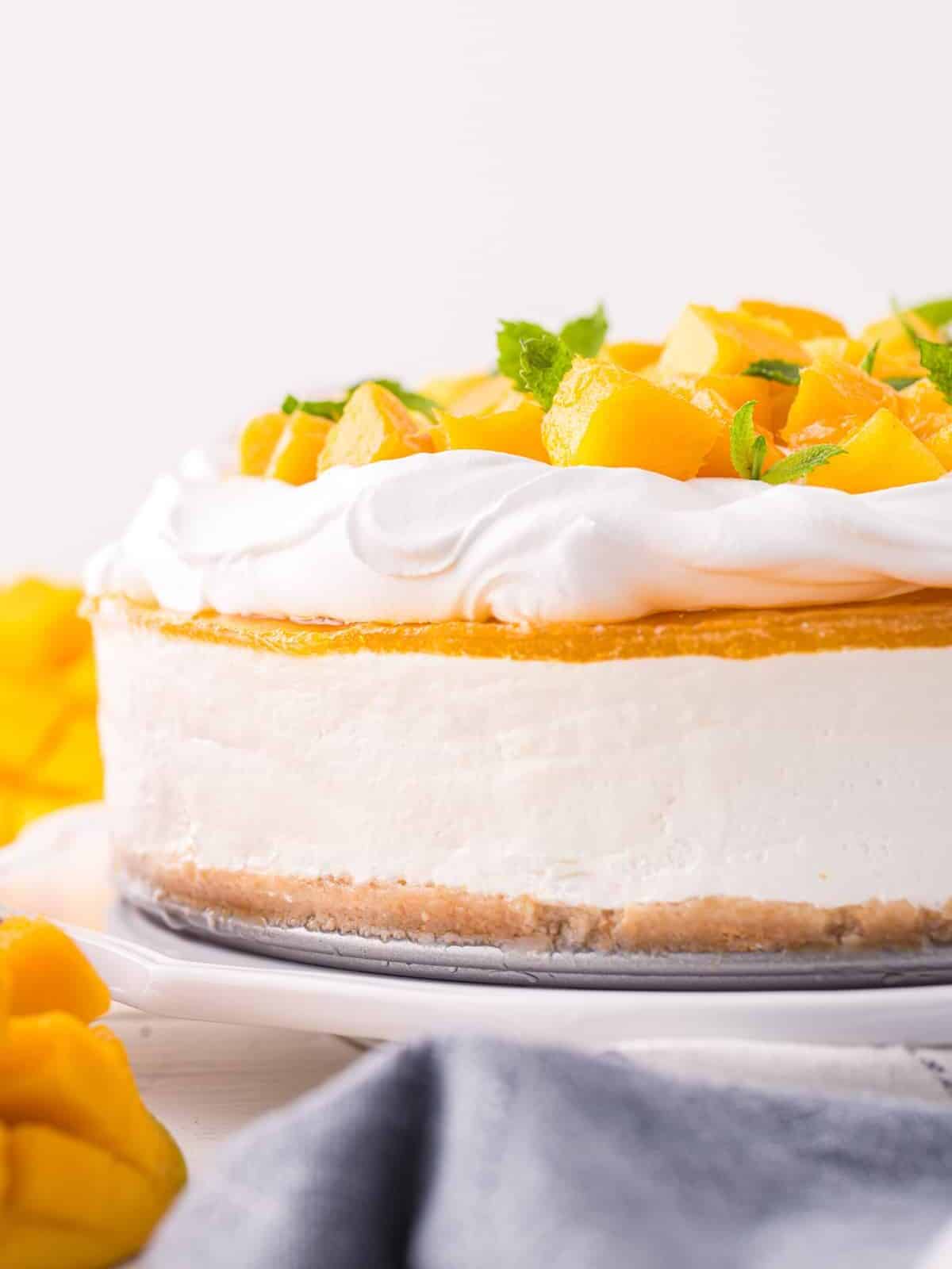 partial side view of mango cheesecake topped with mango chunks and mint on a white cake stand.