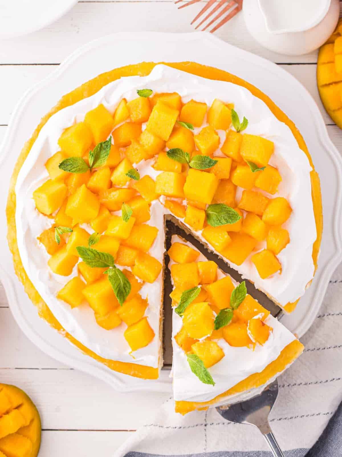 overhead view of a slice being removed from a mango cheesecake topped with mango chunks and mint on a white cake stand.