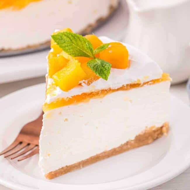 three-quarters view of a slice of mango cheesecake topped with mango chunks and mint on a white plate with a fork.