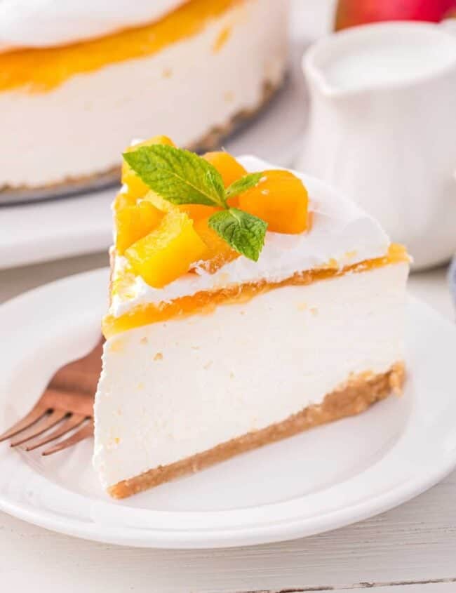 three-quarters view of a slice of mango cheesecake topped with mango chunks and mint on a white plate with a fork.