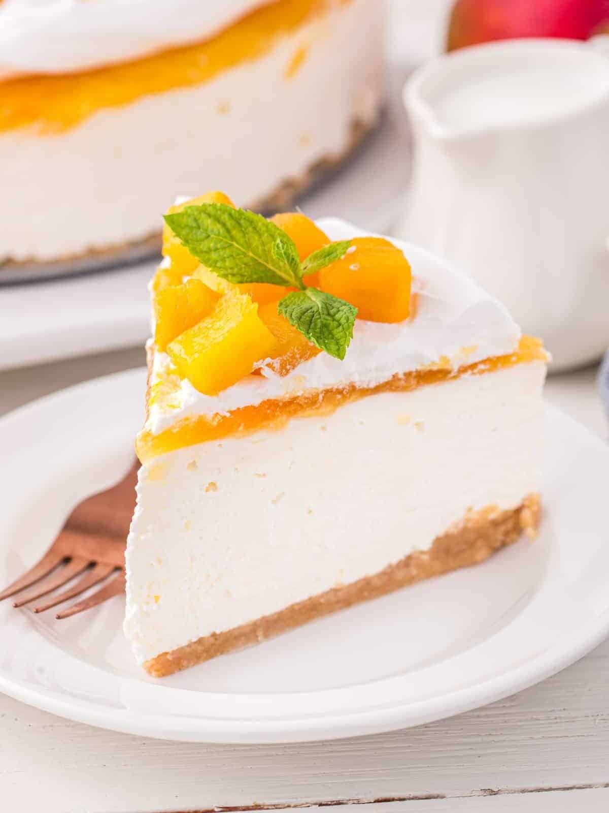 three-quarters view of a slice of mango cheesecake topped with mango chunks and mint on a white plate with a fork.