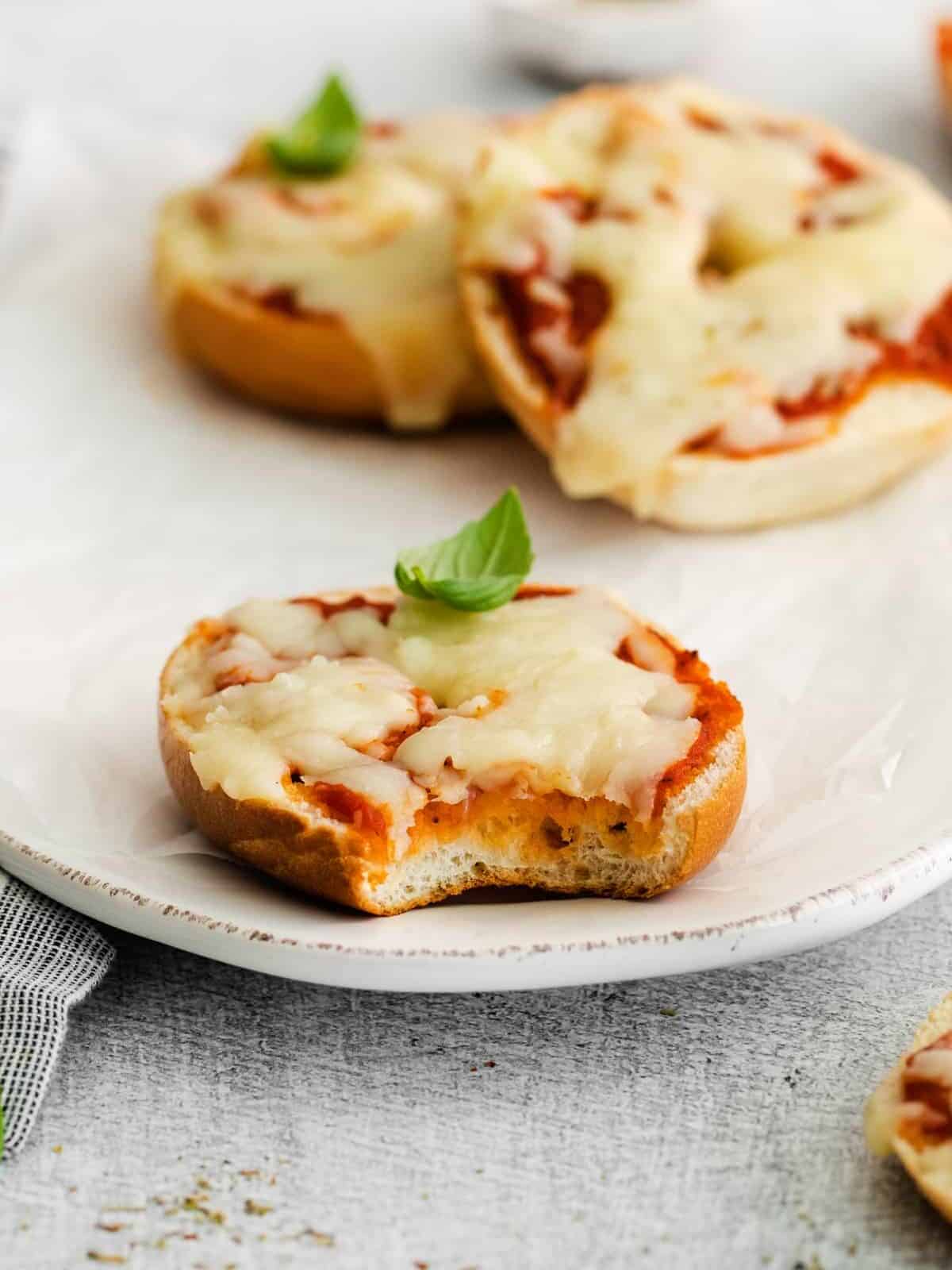 three-quarters view of a bitten homemade pizza bagel on a white oval serving dish in front of 2 more mini pizza bagels.