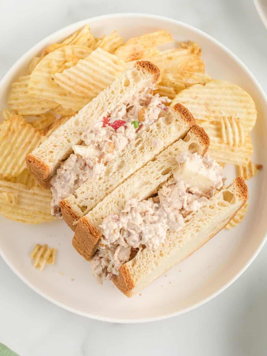 overhead view of a tuna salad sandwich with the halves pointing up to show the filling on a white plate with potato chips.