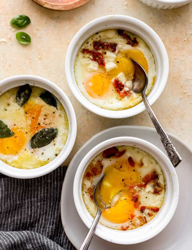 Three bowls of eggs with bacon and basil on a table.