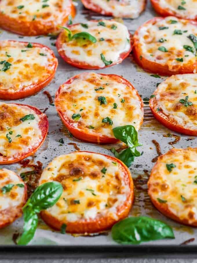 BAKED TOMATOES are a super quick and super easy side dish or appetizer for any occasion! These cheesy Baked Tomatoes with Mozzarella and Parmesan cheese are so simple yet incredibly delicious. They are always a hit when we make them and get eaten right away. These Baked Parmesan Tomatoes are just too tasty and fresh. angled shot of baked tomatoes with cheese and basil