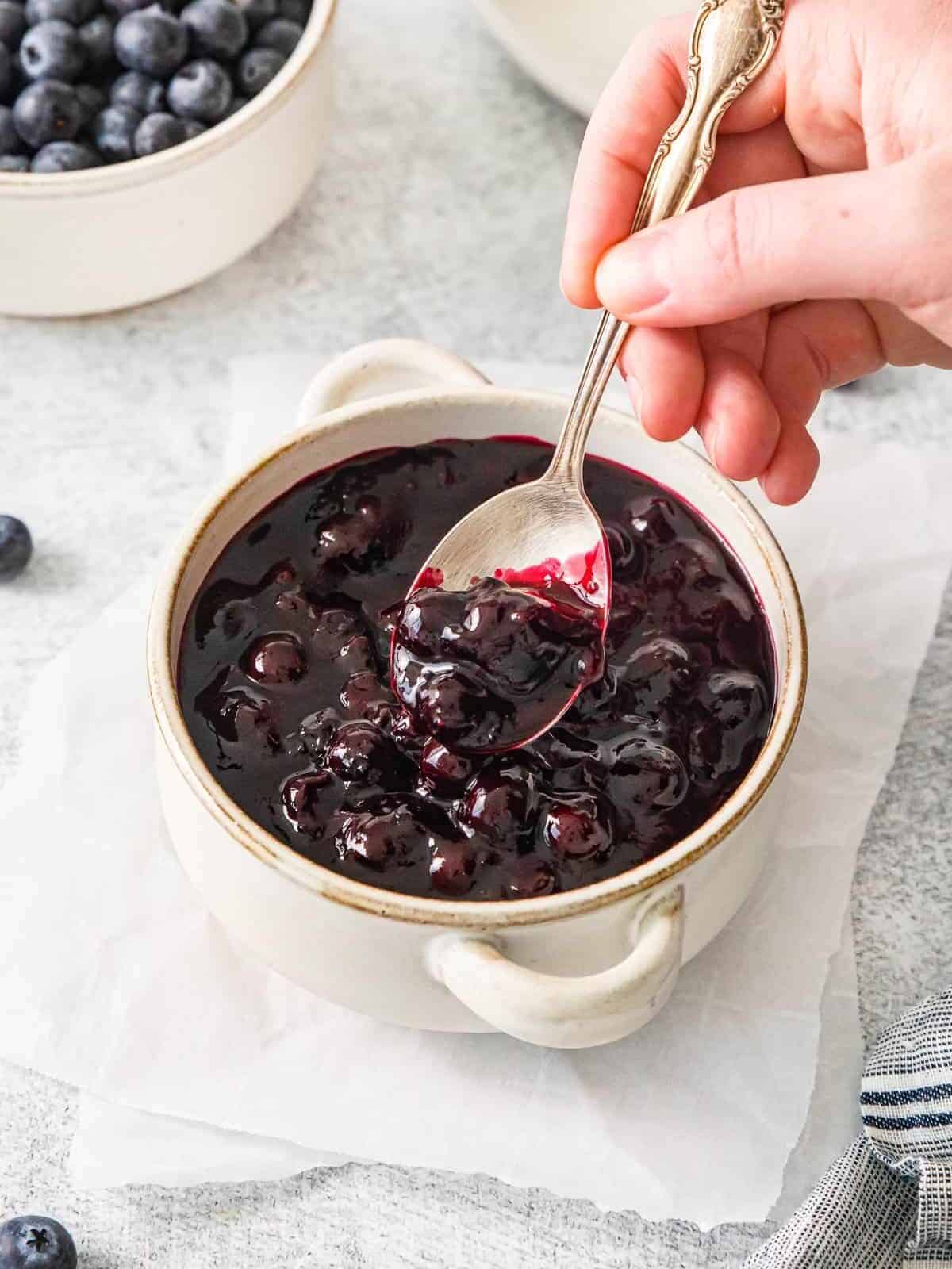 three-quarters view of a spoon scooping fresh blueberry sauce from a small white cocotte.