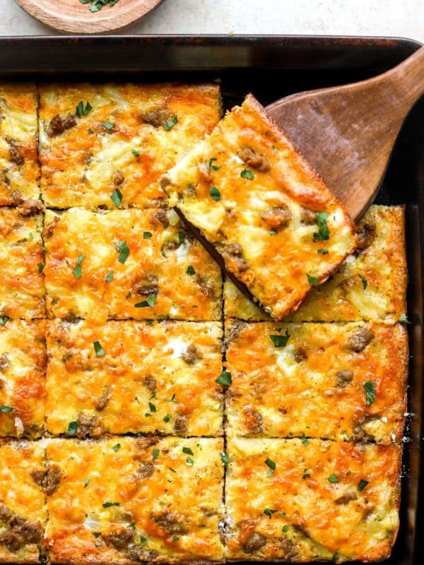 Overhead shot of sheet pan breakfast pizza cut into squares, with one cheesy slice lifted on a wooden spatula.