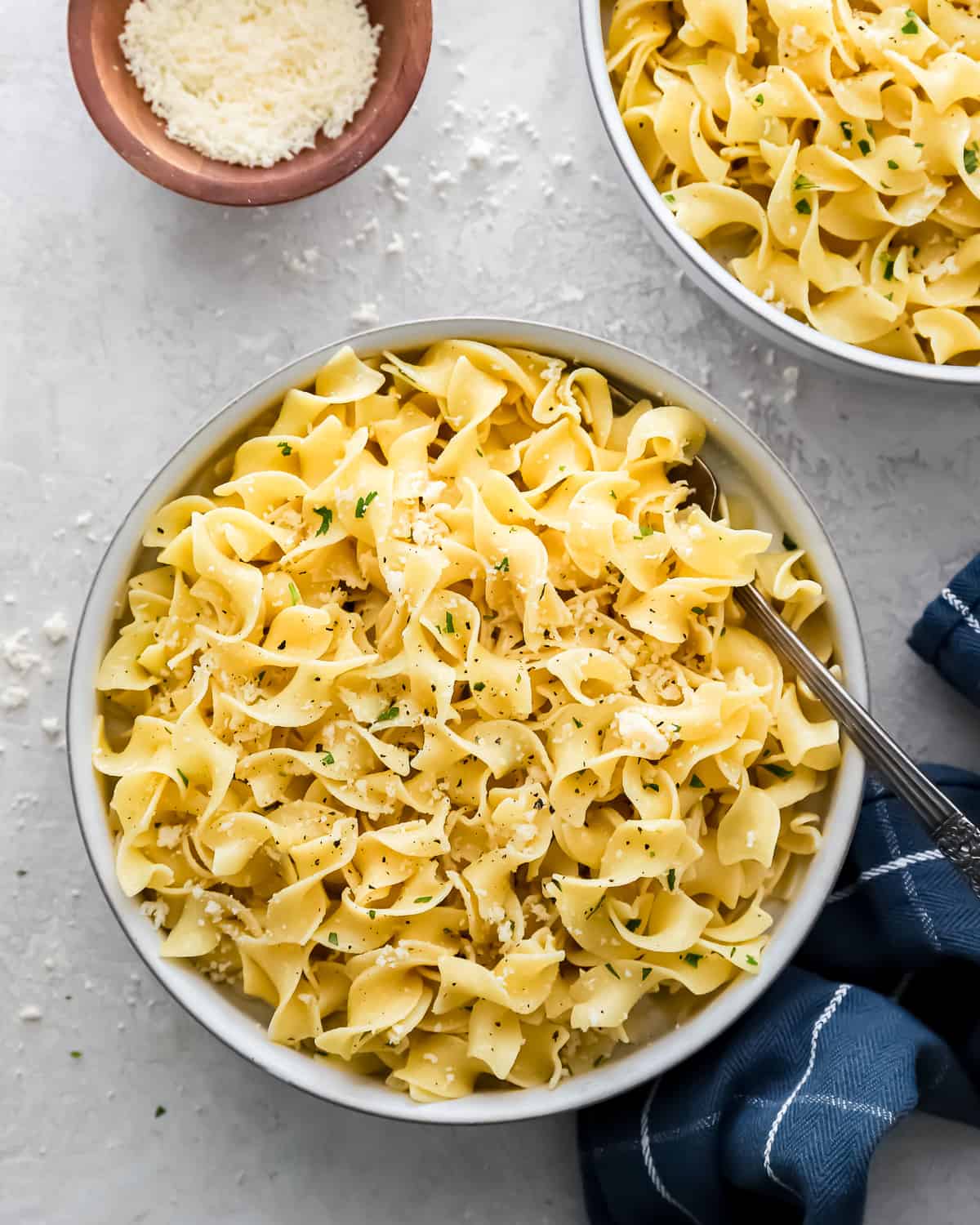 bowls of buttered noodles on a tabletop, next to a small bowl of parmesan cheese.