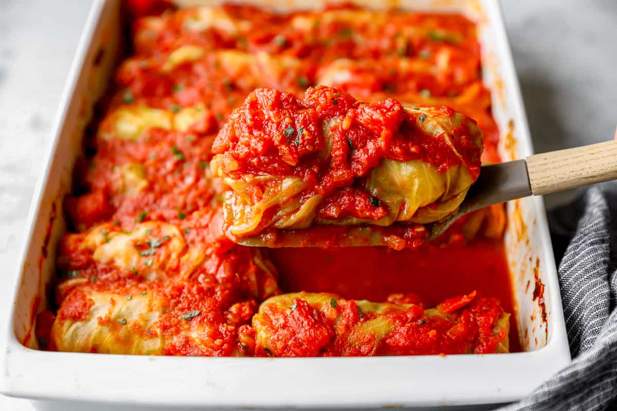 a serving spoon lifting up a cabbage roll above a casserole full of more rolls.