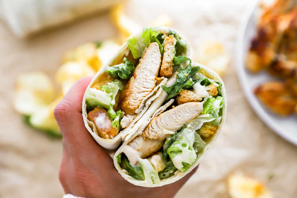 A hand holding a wrap filled with Caesar salad and chicken.