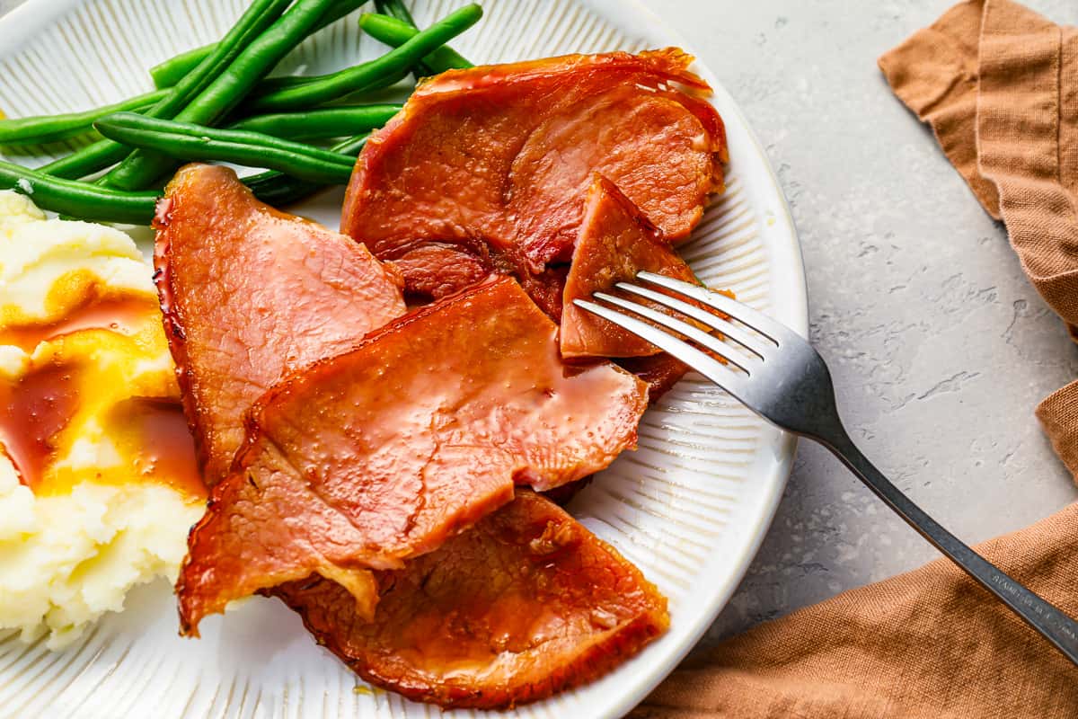 sliced of crockpot ham on a plate with mashed potatoes and green beans.