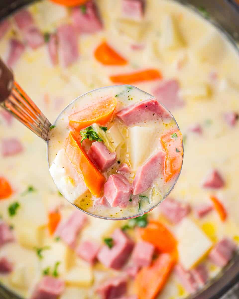 A spoon full of soup with carrots, potatoes and ham.