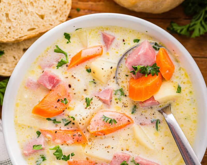 A bowl of soup with carrots, potatoes and ham.