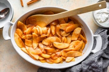 Apples in a white bowl with cinnamon sticks and whipped cream.