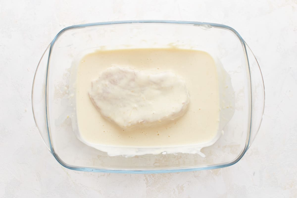 A pork chop in the mayonnaise mixture in a glass bowl.