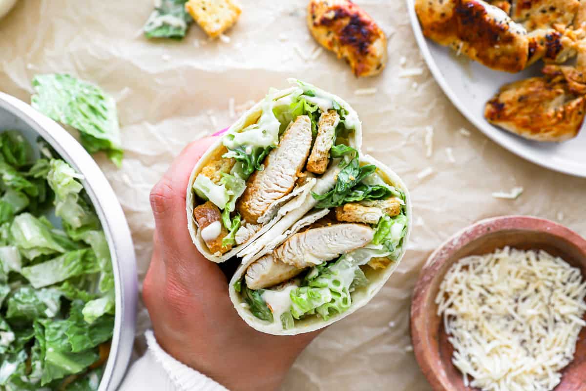 Hand holding the completed chicken Caesar salad wrap to show the pieces of chicken, lettuce, and, croutons inside.