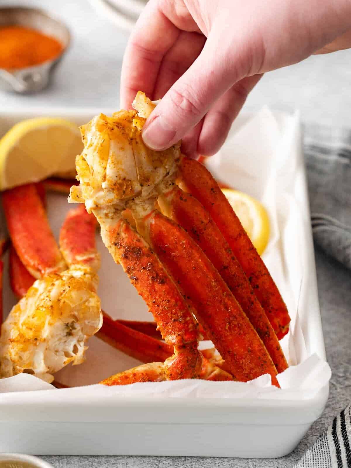 three-quarters view of a hand holding up a cluster of crab legs from a white rectangular plate.