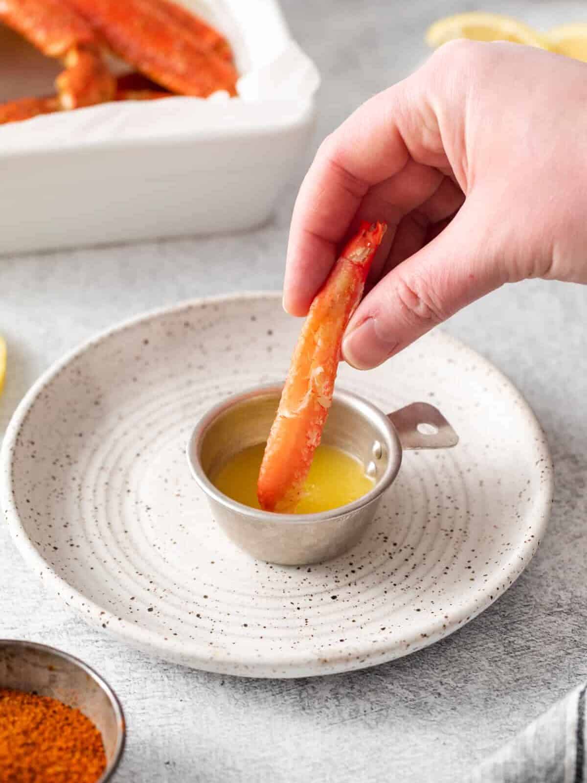 three-quarters view of a hand dipping crab leg meat into a small container of butter on a white plate.