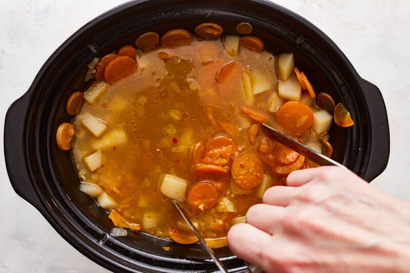 A person stirring carrots and potatoes in a crock pot.