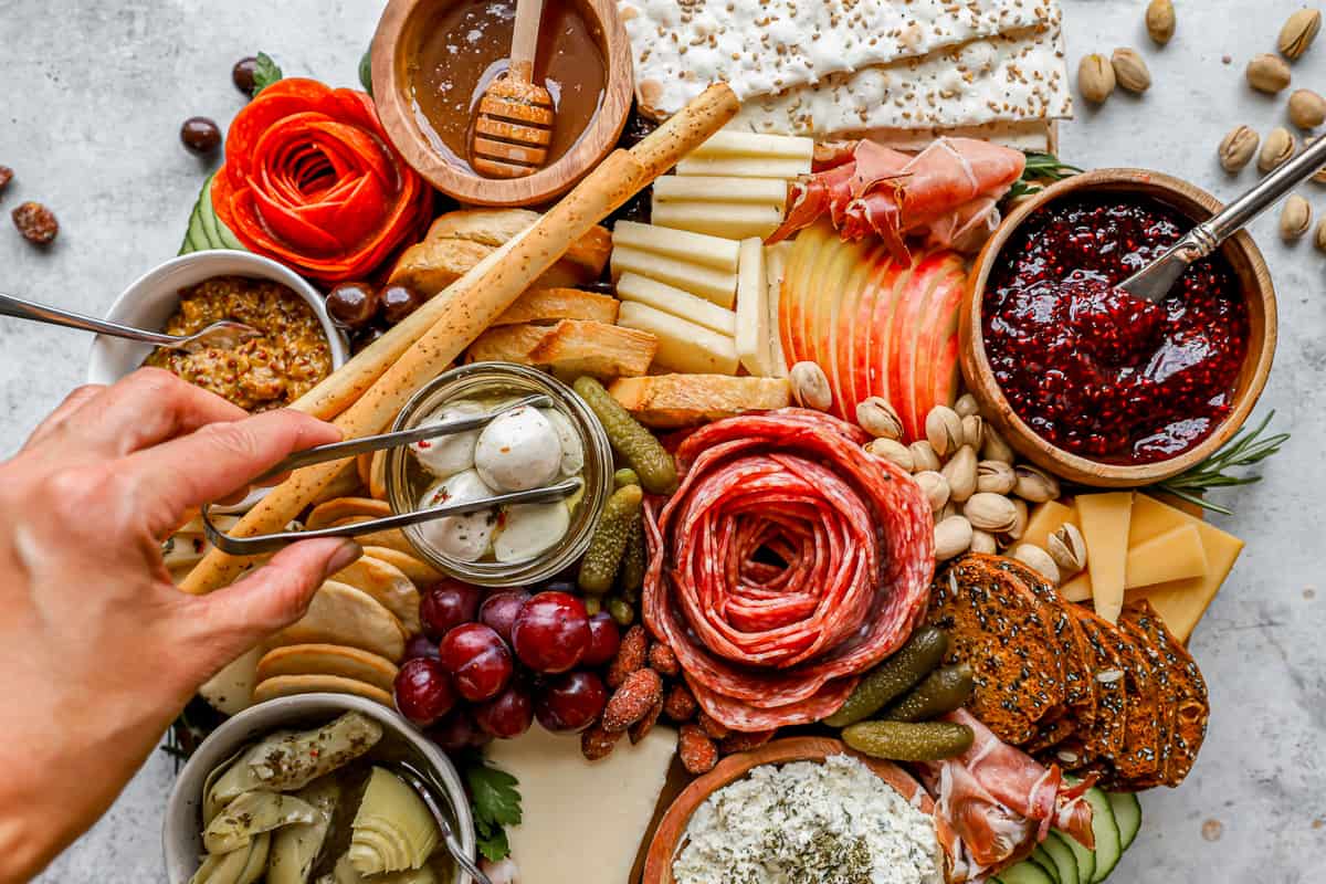 Hands using tongs to grab a mozzarella ball from a charcuterie board.