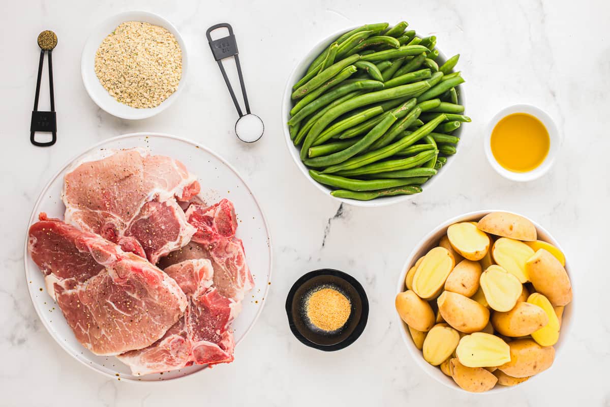Ingredients for this sheet pan pork chops meal, arranged on a marble table. A plate of raw pork chops, a bowl of green beans, a bowl of halved potatoes, and small bowls of seasoning.