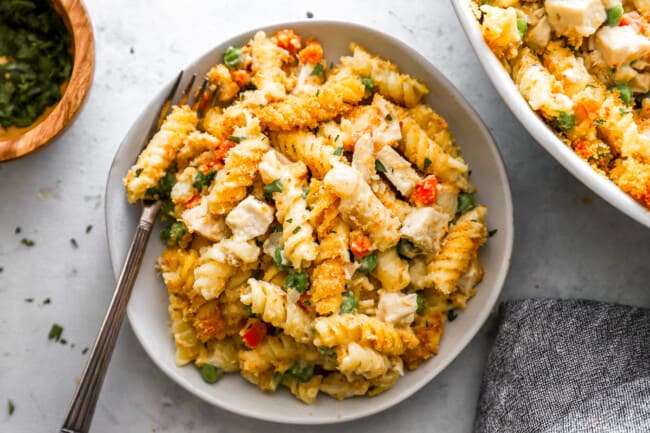 A bowl of creamy rotini pasta mixed with diced vegetables and topped with crumbled cheese. A fork is placed on the side of the dish. There are additional ingredients visible in the background.