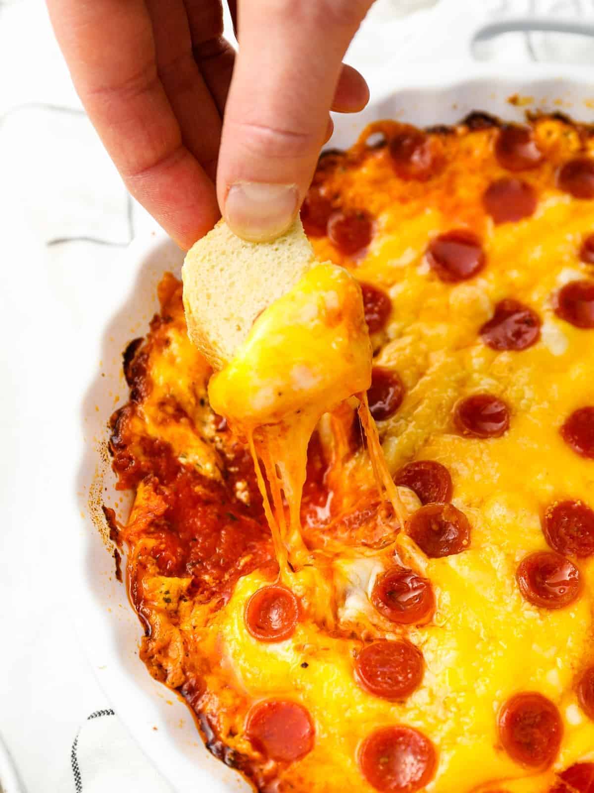 a hand dipping a cube of bread into pepperoni pizza dip in a white round baking dish.