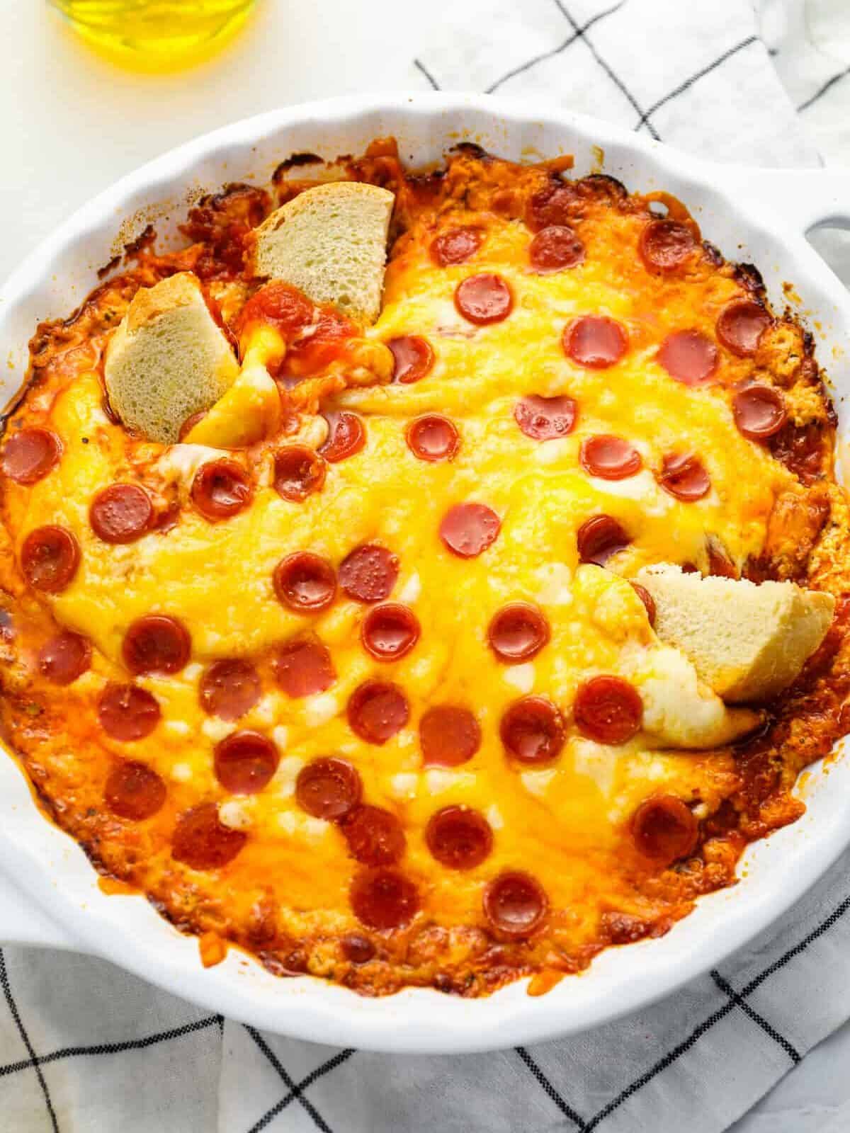 three-quarters view of pizza dip with pepperonis in a white round baking dish with 3 cubes of bread dipped in it.